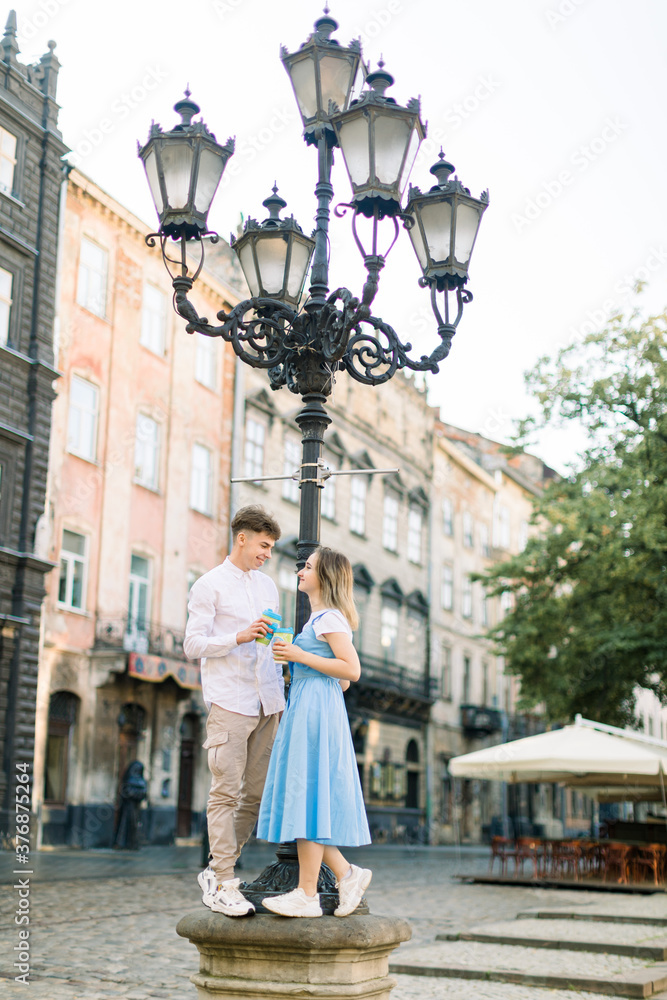 Fototapeta premium Romantic date in old ancient city outdoors. Happy young stylish couple in love with cups of take away coffee, standing under the street light and looking at each other