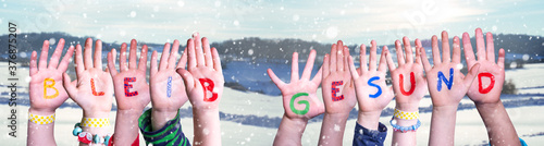 Kids Hands Holding Colorful German Word Bleib Gesund Means Stay Healthy. Snowy Winter Background With Snowflakes