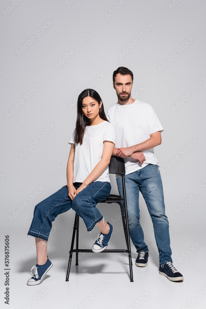 brunette asian woman looking at camera while sitting on chair near stylish boyfriend on grey brunette asian woman looking at camera while sitting on chair near stylish boyfriend on grey