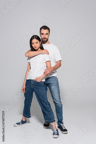 full length view of confident man in white t-shirt and jeans hugging asian girlfriend while posing on grey