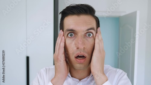 Scared businessman looks into camera with startle at face clutches his head showing fear and surprise. Portrait of shocked man on office background.