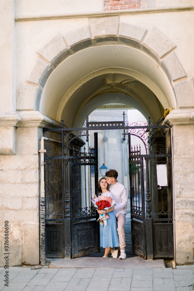City outdoor portrait of happy young couple in love, posing near pod building with beautiful vintage gate. Pretty girl in blue dress holds bouquet of red flowers, handsome guy kisses her