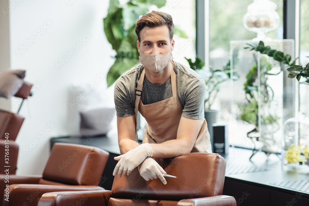 Young male hairdresser in protective mask waiting for clients in salon interior, free space