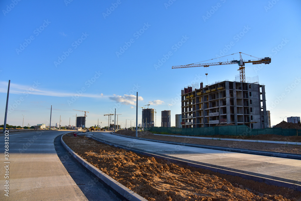 Fototapeta premium View of a large construction site with tall residential buildings and skyscrapers. Tower cranes in action. Road and road junction construction. Laying curbs and paving slabs. Road work on pavement