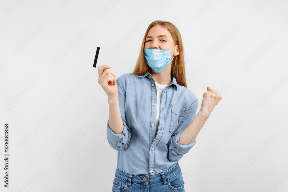 happy beautiful young woman in a medical protective mask on her face shows a winning gesture with a credit card in her hands on a white background happy beautiful young woman in a medical protective mask on her face shows a winning gesture with a credit card in her hands on a white background