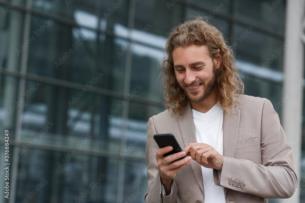 Portrait of handsome curly businessman in casual wear holding smartphone and smiling.Successful manager using mobile phone apps,texting message,browsing internet,looking at phone near business center. Portrait of handsome curly businessman in casual wear holding smartphone and smiling.Successful manager using mobile phone apps,texting message,browsing internet,looking at phone near business center.