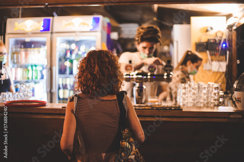 Giovane ragazza con capelli rossi di spalle assiste alla preparazione di una bevanda alcolica in un locale notturno all'aperto photo