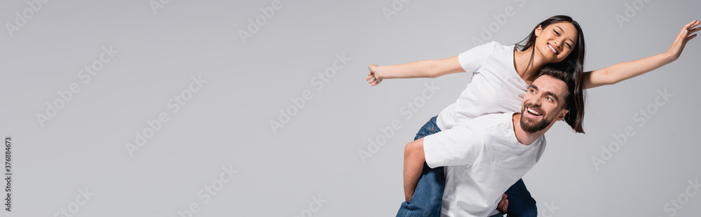 Obraz premium panoramic orientation of excited man piggybacking asian girlfriend imitating flying isolated on grey