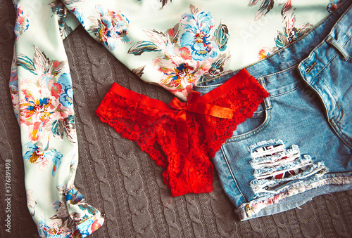 Women's blouse with a floral print on a knitted grey blanket with denim shorts and red lace panties on a bed. © moodysum