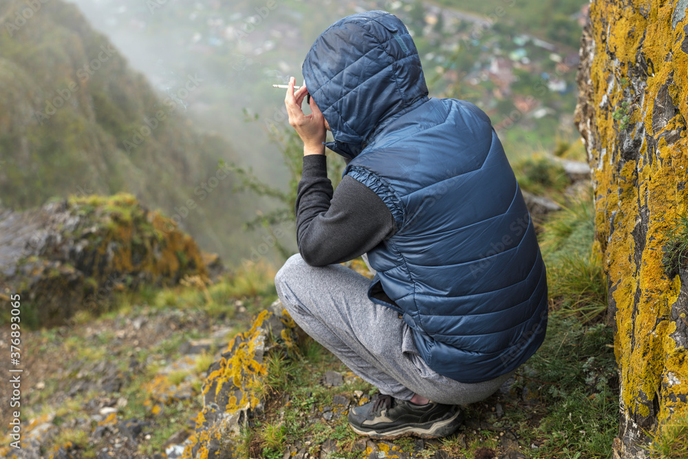A man smokes near a cliff and an abyss in the mountains. Taking pictures while squatting. Back view, hood. Fog, yellow lichen on the rocks. A man smokes near a cliff and an abyss in the mountains. Taking pictures while squatting. Back view, hood. Fog, yellow lichen on the rocks.