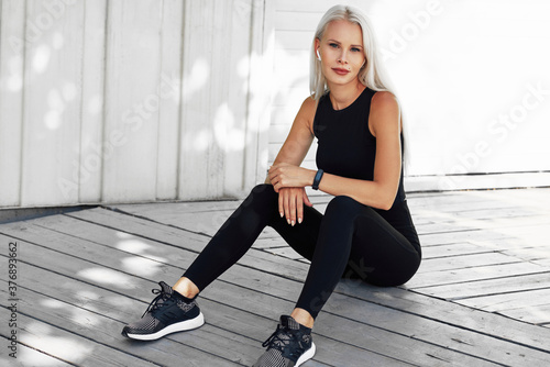 Fototapeta Naklejka Na Ścianę i Meble -  Beautiful blonde female in black sportswear resting after workout exercises outdoors. Fitness girl takes a rest after running in the morning sitting agaisnt white wall outside.