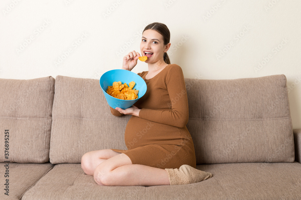 Pregnant woman sitting on the sofa enjoys eating potato chips from a bowl at home. Feeling hungry during pregnancy concept Pregnant woman sitting on the sofa enjoys eating potato chips from a bowl at home. Feeling hungry during pregnancy concept