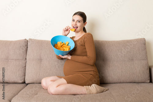 Pregnant woman sitting on the sofa enjoys eating potato chips from a bowl at home. Feeling hungry during pregnancy concept