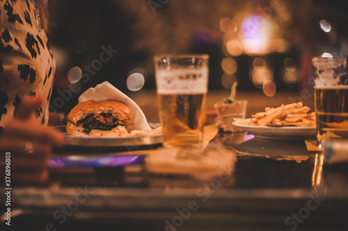 Dettaglio primo piano panino hamburger, patatine fritte e bicchiere di birra fresca su tavolo in legno di un locale notturno all'aperto photo