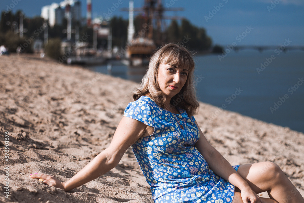 Young blonde woman in blue dress sits on the sand in the park