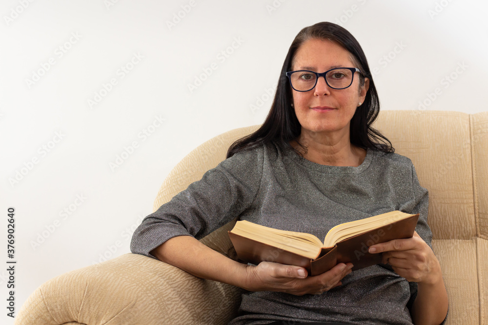 Christian Middle-aged woman with the Bible in her hands seated in cream armchair and looking at the camera. Christian Middle-aged woman with the Bible in her hands seated in cream armchair and looking at the camera.