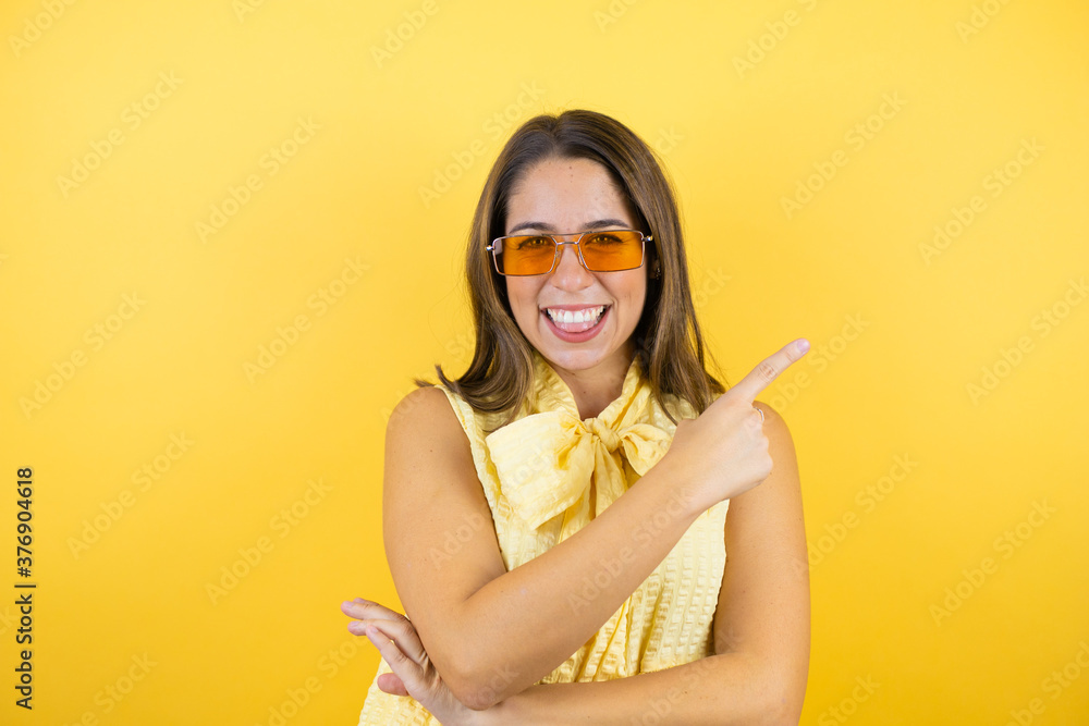 Young beautiful woman wearing sunglasses over isolated yellow background smiling happy pointing with hand and finger to the side Young beautiful woman wearing sunglasses over isolated yellow background smiling happy pointing with hand and finger to the side