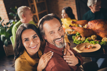Closeup photo of full family gathering bearded husband wife cuddle beaming smiling hold hands finish eat meal sit served dinner big table turkey generation in evening living room indoors