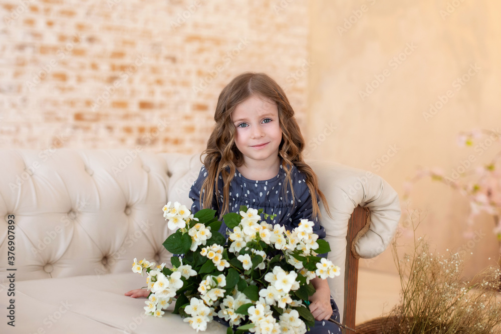 Smiling little curly girl with bouquet of jasmine flowers sits on couch in living room. Childhood. Celebration. Mothers Day. Child with bouquet of flowers for mom. Girl with flowers for birthday. 