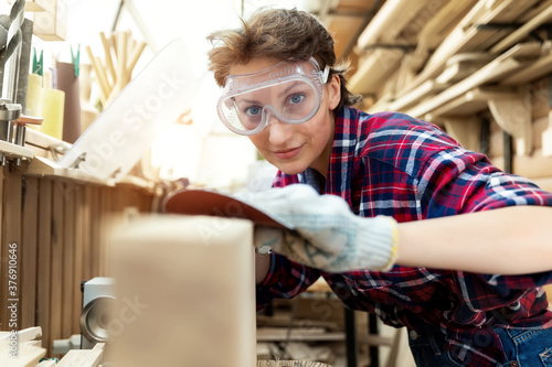 Young attractive 30-40 professional female carpenter looking grinding raw wood with sandpaper in carpentry diy workshop. Feminine women equality concept. Adult girl with male hobby at workbench