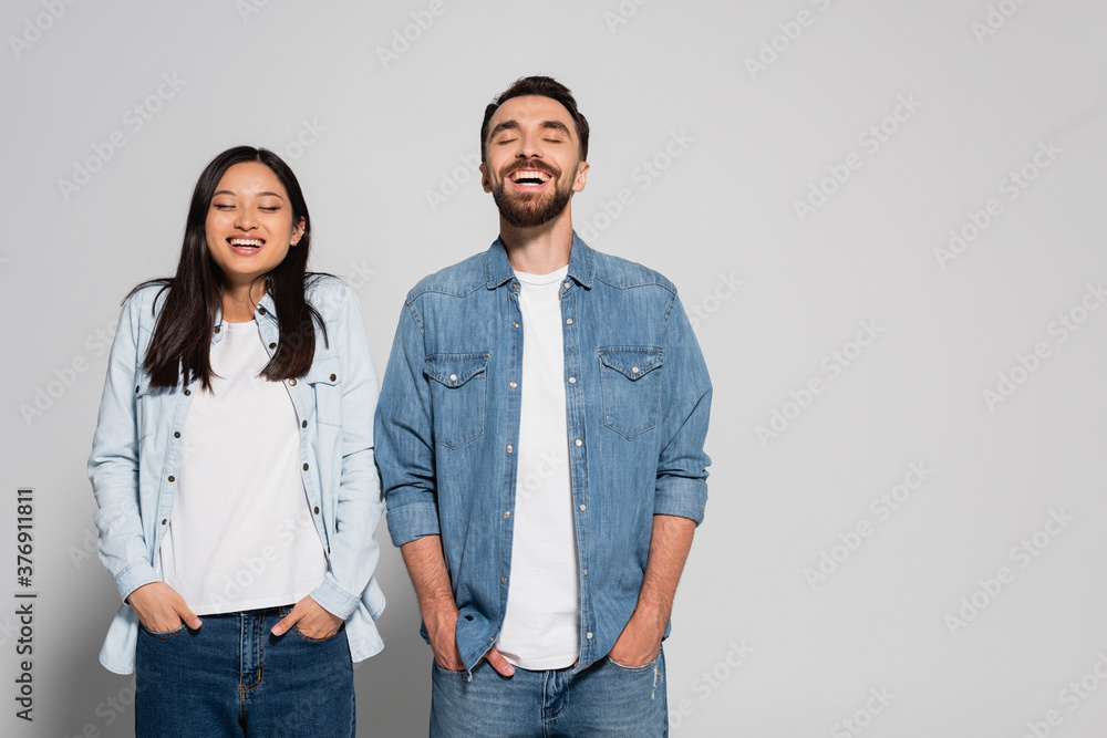 young interracial couple in denim clothes holding hands in pockets while standing with closed eyes on grey young interracial couple in denim clothes holding hands in pockets while standing with closed eyes on grey