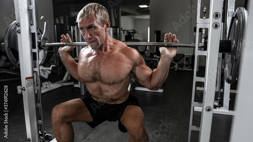 Muscular adult brutal man doing barbell squats in the gym. Portrait of caucasian authentic bodybuilder doing workout exercises