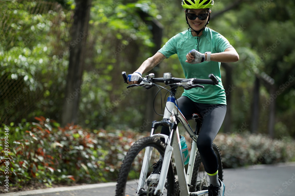 Naklejka premium Woman cyclist looking at her smartwatch while riding bike outdoors on sunny day