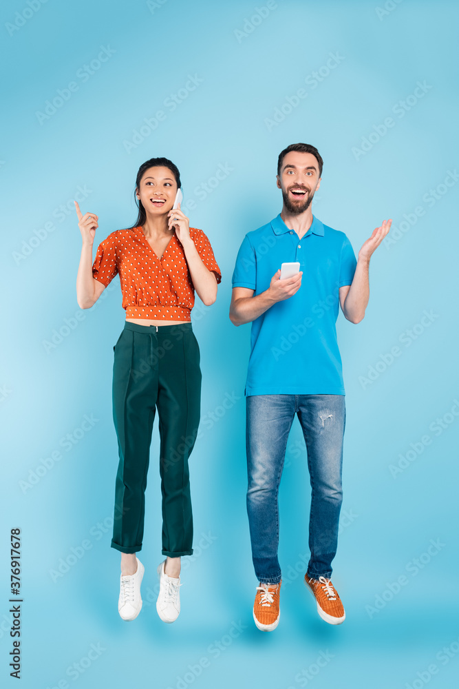 brunette asian woman talking on smartphone and pointing with finger near excited man levitating on blue