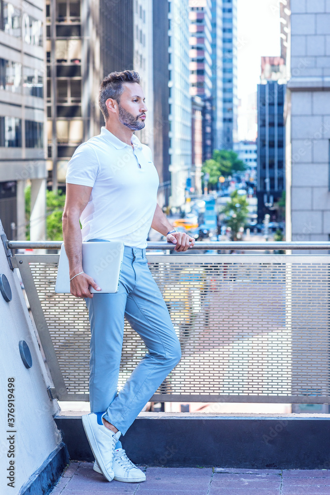 Young Serious European Businessman with beard, little gray hair, wearing white shirt, light blue pants, white sneakers, holding laptop computer, standing by railing on balcony in New York City..