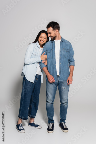 full length view of young asian woman with closed eyes leaning on stylish boyfriend on grey