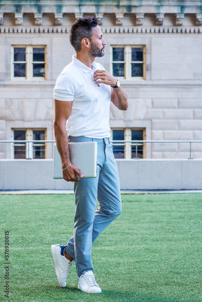 Young European Businessman with beard, little gray hair, wearing white short sleeve shirt, light blue pants, white sneakers, holding laptop computer, standing on green lawn outside in New York City..