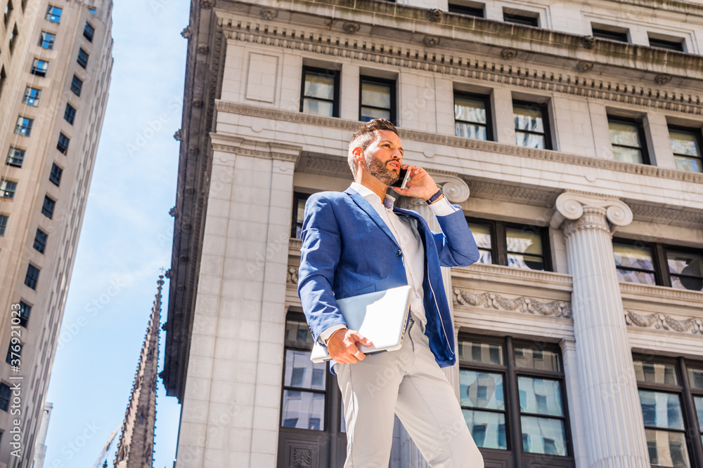 Young European Businessman with beard traveling, working in New York City, wearing blue blazer, gray pants, holding laptop computer, standing outside old office building, calling on cell phone..