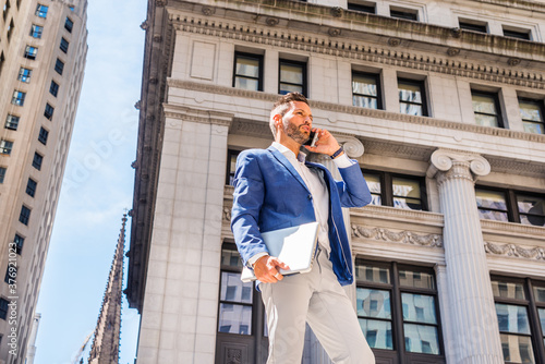 Obraz na płótnie Young European Businessman with beard traveling, working in New York City, wearing blue blazer, gray pants, holding laptop computer, standing outside old office building, calling on cell phone.. w salonie