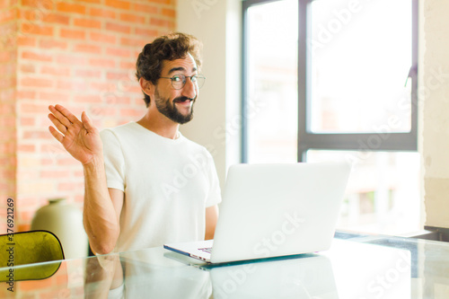 young bearded man with a laptop smiling happily and cheerfully, waving hand, welcoming and greeting you, or saying goodbye photo