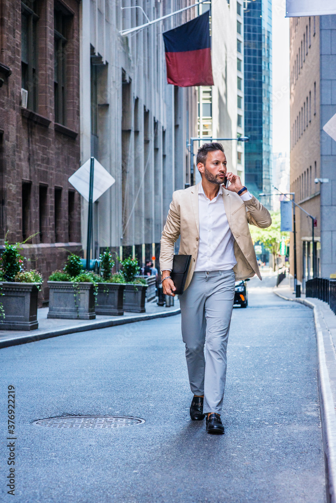 European Businessman traveling, working in New York City, with beard, little gray hair, wearing beige blazer, gray pants, leather shoes, holding briefcase, walking on street, talking on cell phone..