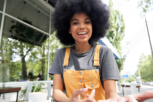 Young African American mixed race hipster vlogger influencer woman with Afro hair looks at webcam talks to camera sits at outdoor cafe table video calling, recording blog, headshot portrait.