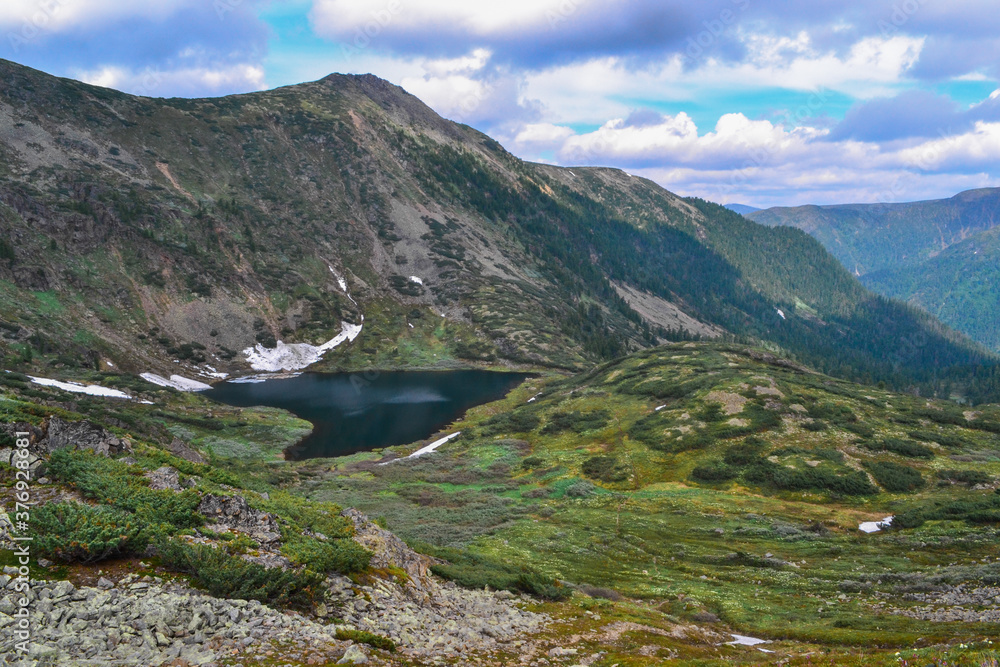 view of mountain lake in form of heart among snow, green mountain ranges overgrown with trees, Chersky peak, clouds