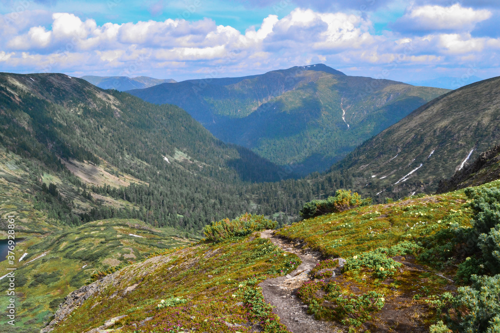 trail in blooming green meadow with flowers among blue Baikal mountains .ridge, blue sky, clouds