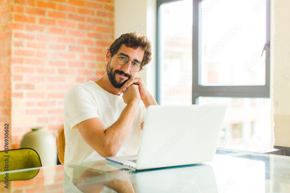 young bearded man with a laptop feeling in love and looking cute, adorable and happy, smiling romantically with hands next to face - obrazy, fototapety, plakaty 