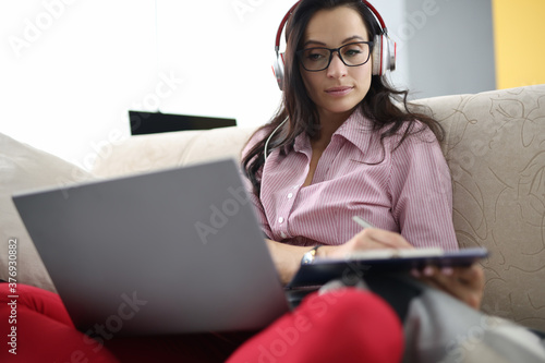 Quadro em tela Woman in headphones with clipboard in her hands sits on couch with laptop