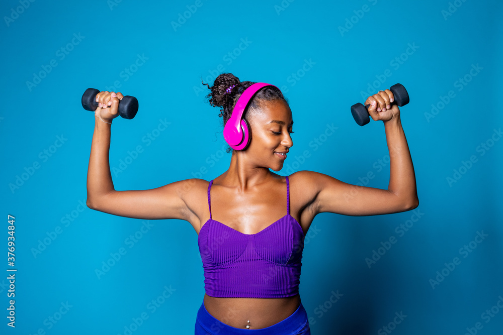 Fit young black woman weight lifting on blue background listening music - Isolated diverse black female workout with weights wearing headphones - fitness, training, sport concept Fit young black woman weight lifting on blue background listening music - Isolated diverse black female workout with weights wearing headphones - fitness, training, sport concept