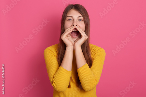 Cheerful funny young woman wearing yellow sweater screaming with hand gesture near mouth isolated over pink wall, lady with long hair, screams loudly.