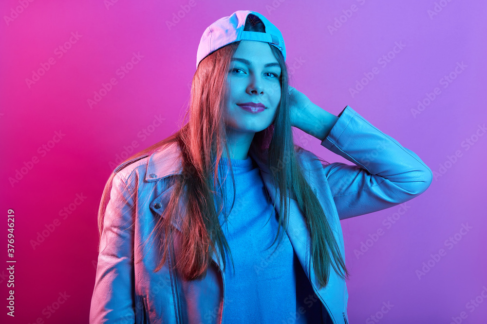 Beautiful girl portrait wearing basket cap and leather jacket, looking directly at camera, keeping hand on head, has long hair isolated over pink neon wall. Beautiful girl portrait wearing basket cap and leather jacket, looking directly at camera, keeping hand on head, has long hair isolated over pink neon wall.