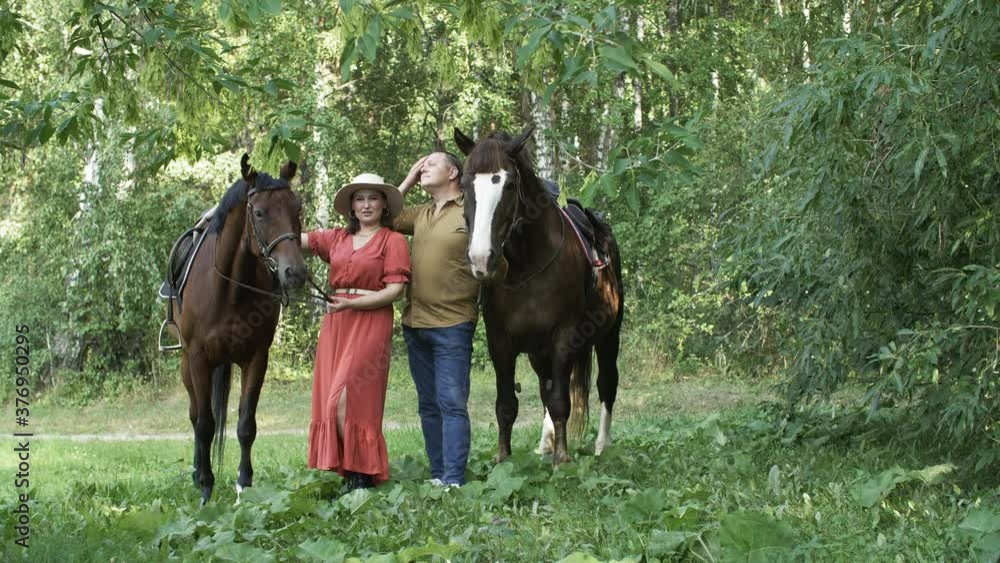 Beautiful middle aged couple standing with horses on green meadow. Man and woman holding bridles and standing between majestic brown horses in countryside Beautiful middle aged couple standing with horses on green meadow. Man and woman holding bridles and standing between majestic brown horses in countryside