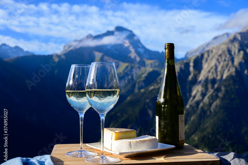 Tasty cheese and wine from Savoy region in France,  tomme and reblochon de savoie cheeses and glass of white wine served outdoor with Alpine mountains peaks on background photo