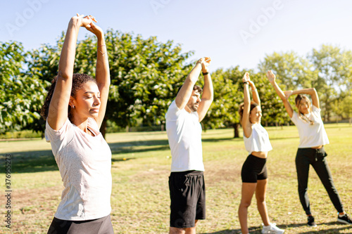 Beautiful young curly hispanic woman teach training with closed eyes together with her three friends in the nature - Cute latina athletics instructor stretching her arms with closed eyes in the park