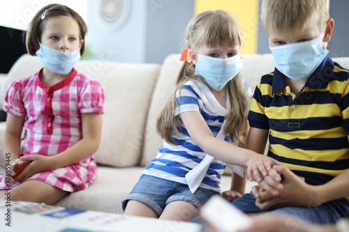 Pensive children are sitting on couch wearing protective medical masks and holding board playing cards. Safe communication in a team in coronavirus concept