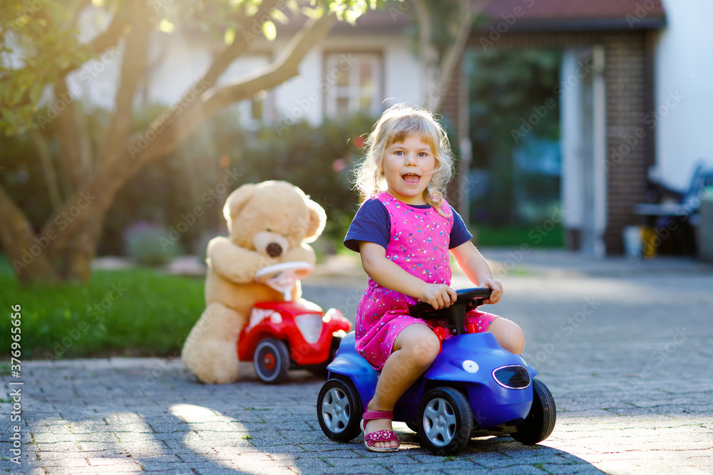 Little adorable toddler girl driving toy car and having fun with playing with plush toy bear, outdoors. Gorgeous happy healthy child enjoying warm summer day. Smiling stunning kid in gaden Little adorable toddler girl driving toy car and having fun with playing with plush toy bear, outdoors. Gorgeous happy healthy child enjoying warm summer day. Smiling stunning kid in gaden