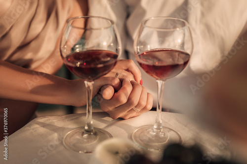 Close up shot of the bride and groom hands. Young couple have a date, celebrating the honeymoon and drinking red wine, spending time together. Husband and wife with wedding rings. Valentines day.