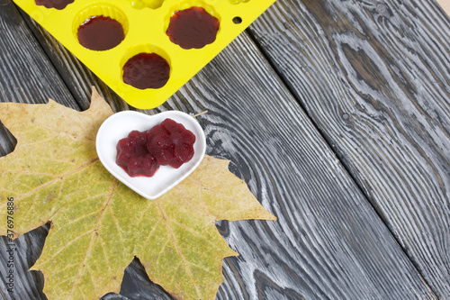 Saucer with cherry marmalade. A silicone mold with marmalade is visible. Dried maple leaves all around. On pine planks painted black and white.